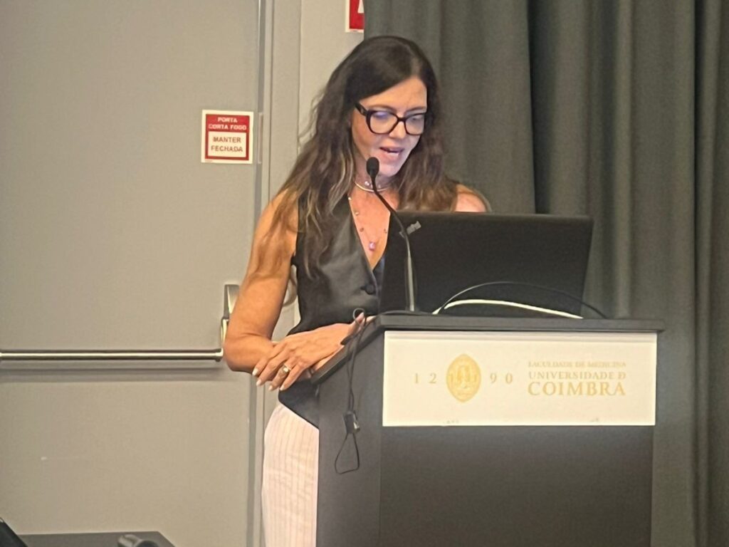Mulher usando óculos fala ao microfone enquanto apresenta slides em um púlpito da Faculdade de Medicina da Universidade de Coimbra.