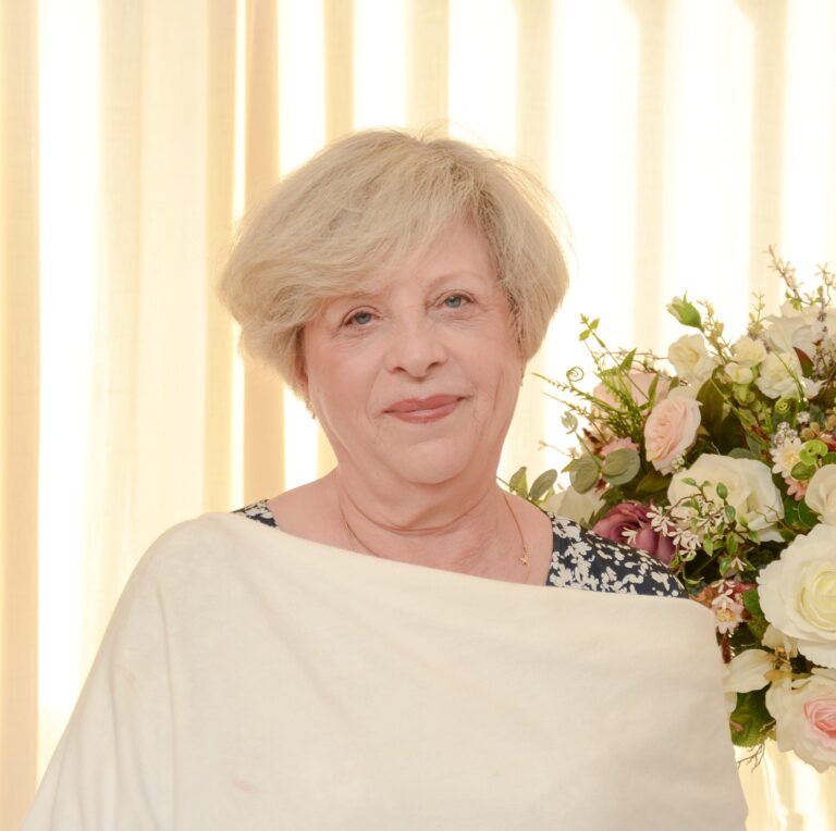 Mulher sorridente de cabelos curtos e claros, usando uma blusa clara sobre um vestido estampado, posa em frente a uma cortina bege, ao lado de um arranjo de flores em tons claros.