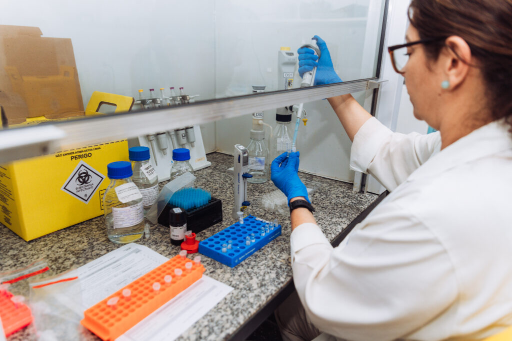 Pessoa vestindo jaleco e luvas azuis trabalha em bancada de laboratório, manuseando uma micropipeta dentro de uma cabine de segurança. Sobre a superfície há tubos de ensaio coloridos, frascos rotulados, materiais de análise e uma caixa amarela de descarte de resíduos biológicos.