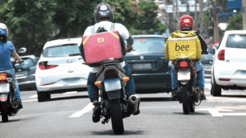 Três motociclistas circulam por uma rua movimentada durante o dia. Dois deles carregam mochilas de entrega nas costas, uma vermelha e outra amarela com o nome “bee”. Há carros à frente e ao lado, e árvores e construções ao fundo.