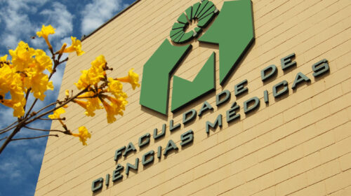 Fachada da Faculdade de Ciências Médicas da Unicamp, com o logotipo verde em destaque sobre parede amarelada. No canto esquerdo da imagem, galhos de ipê-amarelo florescem sob um céu azul com nuvens.