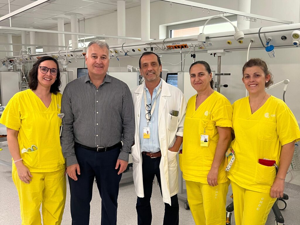 Cinco profissionais da saúde posam em uma sala de atendimento hospitalar; quatro deles vestem uniformes amarelos e um homem ao centro veste camisa listrada, com outro homem de jaleco branco ao lado.