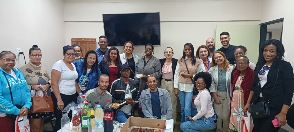 “Grupo de aproximadamente vinte pessoas reunidas em uma sala para um momento de confraternização. Estão posicionadas em pé e sentadas ao redor de uma mesa com bebidas e bolo, sorrindo para a foto.”
