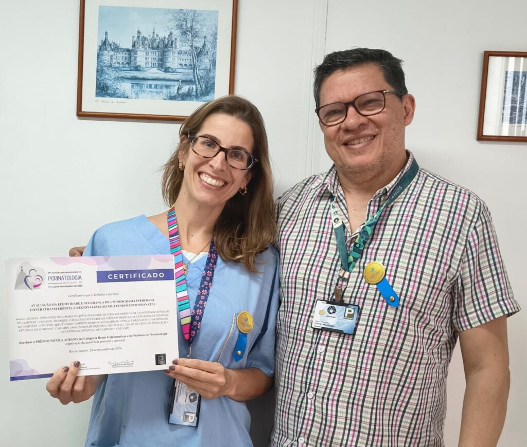 "Duas pessoas sorriem para a foto em um ambiente interno. A mulher à esquerda, vestindo avental azul e crachá colorido, segura um certificado do Congresso Brasileiro de Perinatologia. Ao lado, um homem com camisa xadrez e crachá também sorri. Ambos usam pequenos bottons amarelos presos ao crachá."