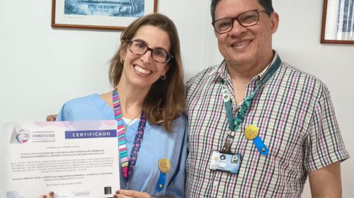 "Duas pessoas sorriem para a foto em um ambiente interno. A mulher à esquerda, vestindo avental azul e crachá colorido, segura um certificado do Congresso Brasileiro de Perinatologia. Ao lado, um homem com camisa xadrez e crachá também sorri. Ambos usam pequenos bottons amarelos presos ao crachá."