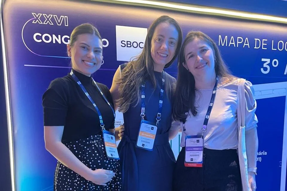 Três mulheres sorrindo e posando para a foto durante o XXVI Congresso da SBOC. Elas usam crachás do evento e estão posicionadas diante de um painel iluminado com informações do congresso.