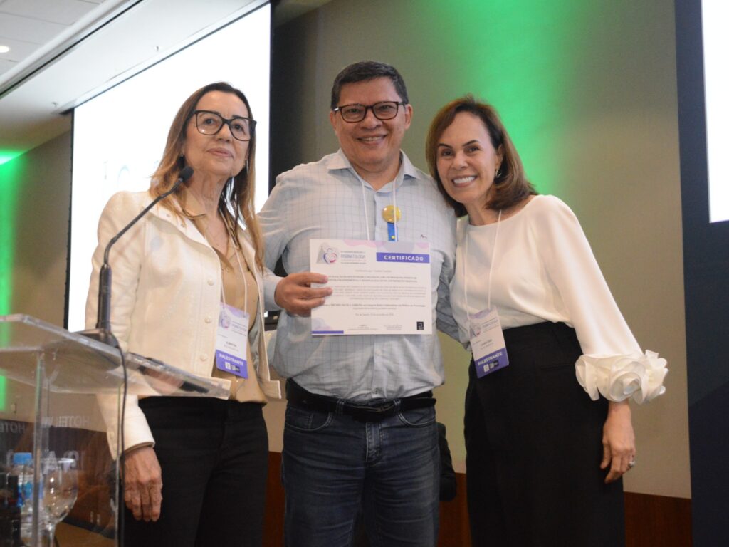 "Três pessoas posam sorrindo durante um evento. À esquerda, uma mulher fala ao microfone em um púlpito. Ao centro, um homem segura um certificado do Congresso Brasileiro de Perinatologia. À direita, outra mulher também sorri para a câmera. Todos usam crachás de identificação."