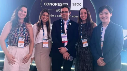 Grupo de cinco participantes do XXVI Congresso da SBOC posando para foto em frente ao painel principal do evento. Quatro mulheres e um homem usam crachás e sorriem enquanto se posicionam lado a lado.
