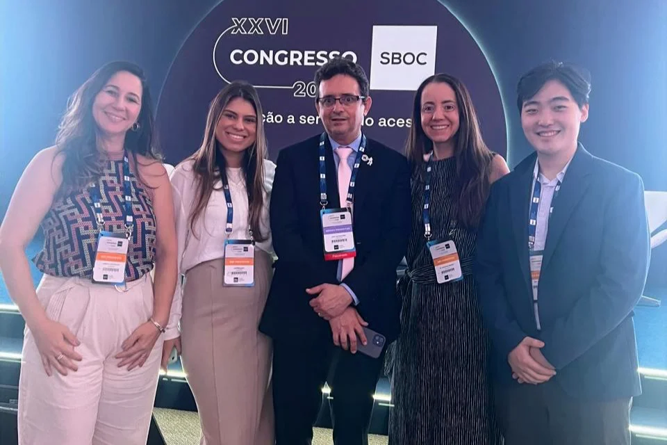 Grupo de cinco participantes do XXVI Congresso da SBOC posando para foto em frente ao painel principal do evento. Quatro mulheres e um homem usam crachás e sorriem enquanto se posicionam lado a lado.