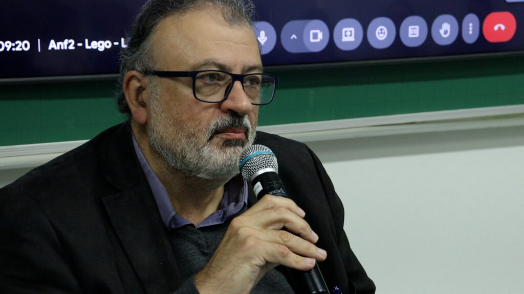 Homem fala ao microfone durante evento em sala de aula, com quadro verde e tela de videoconferência ao fundo.