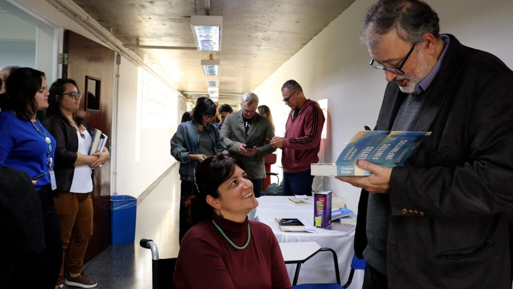 Homem autografa um livro para uma mulher sentada, enquanto outras pessoas aguardam em fila em um corredor iluminado.
