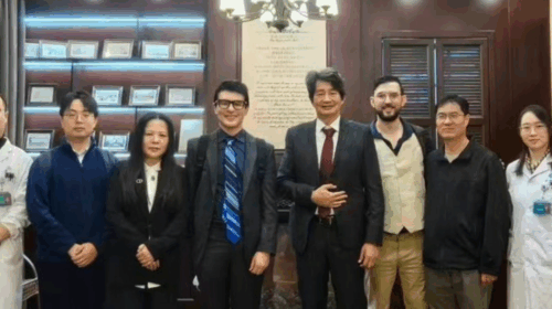 “Grupo de nove pessoas posando para foto em um ambiente interno elegante, com estantes de madeira ao fundo contendo certificados e molduras. Entre elas, há profissionais usando jalecos brancos e outras pessoas vestindo roupas formais. Todos estão alinhados lado a lado, olhando para a câmera.”