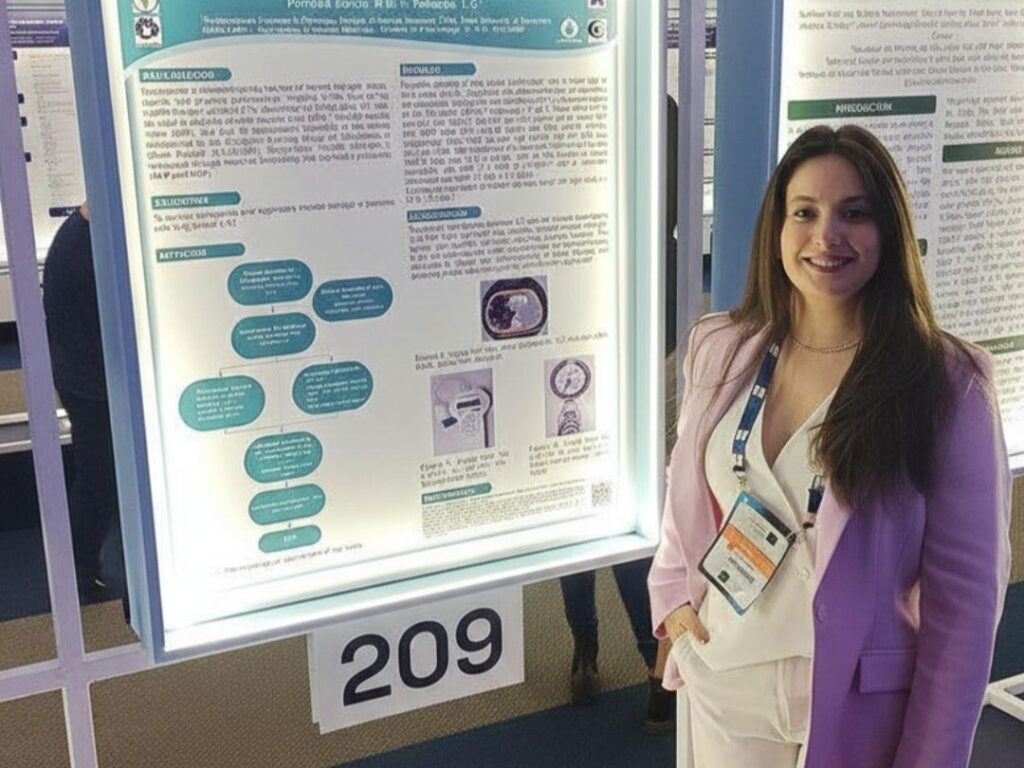 Mulher sorrindo ao lado de um painel iluminado que exibe seu pôster científico numerado como 209, durante apresentação em congresso. Ela veste blazer lilás e roupa clara, com crachá do evento pendurado no pescoço.