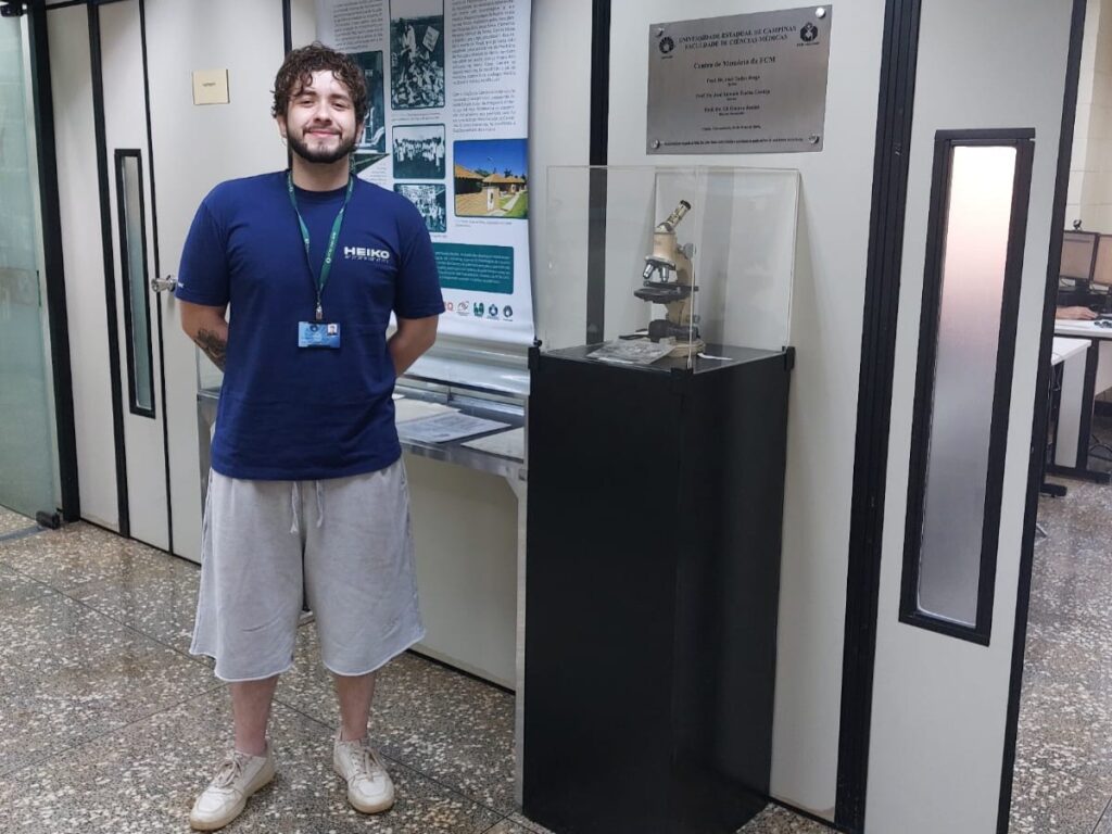 Um homem jovem está em pé em um corredor interno, sorrindo para a câmera. Ele veste camiseta azul com crachá no pescoço, shorts claros e tênis. Atrás dele há um painel com informações históricas e, ao lado, uma vitrine preta com um microscópio antigo exposto. O ambiente parece ser uma área institucional ou de pesquisa, com portas e divisórias de vidro ao fundo.