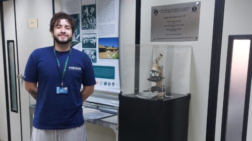 Um homem jovem está em pé em um corredor interno, sorrindo para a câmera. Ele veste camiseta azul com crachá no pescoço, shorts claros e tênis. Atrás dele há um painel com informações históricas e, ao lado, uma vitrine preta com um microscópio antigo exposto. O ambiente parece ser uma área institucional ou de pesquisa, com portas e divisórias de vidro ao fundo.