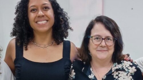 Duas mulheres posam juntas e sorriem. A mulher à esquerda tem cabelos cacheados, veste uma blusa preta de alças largas e usa colares delicados. A mulher à direita tem cabelos lisos na altura dos ombros, usa óculos e veste uma blusa escura estampada com flores claras. Elas estão em um ambiente interno com fundo claro.