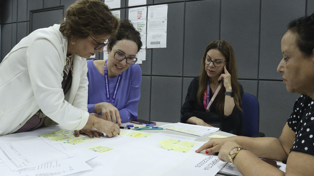 Quatro mulheres sentadas em torno de uma mesa participam de uma atividade em grupo durante o Planejamento Estratégico do ProDDocS da FCM Unicamp. Elas analisam anotações em papéis e post-its espalhados sobre a mesa, em um ambiente de trabalho colaborativo. Ao fundo, parede cinza com documentos afixados.
