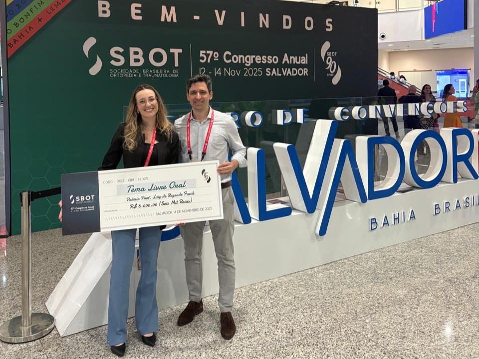 A Dra. Érica Blenda e seu orientador, Professor Guilherme Mouraria, posam sorridentes segurando um cheque simbólico gigante. O cheque refere-se ao "Prêmio Prof. Luiz de Rezende Puech" na categoria "Tema Livre Oral", no valor de R$ 6.000,00. Eles estão no saguão do evento, em frente a um letreiro volumétrico branco escrito "SALVADOR" e um banner do 57º Congresso Anual da SBOT.