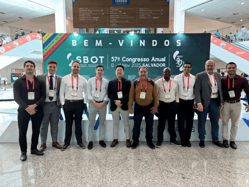 Foto posada de um grupo de dez homens, incluindo médicos residentes, um acadêmico e os professores Guilherme Mouraria e Maurício Etchebehere. Eles estão de pé, lado a lado, sorrindo e usando crachás vermelhos do evento. Ao fundo, um grande painel verde traz a mensagem "BEM-VINDOS" e as informações do "SBOT - 57º Congresso Anual", realizado em Salvador em novembro de 2025.
