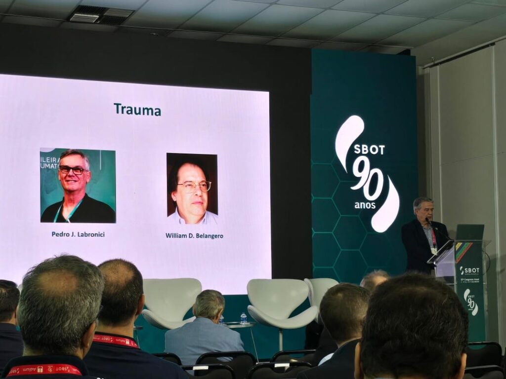 Foto de um auditório focada no telão de projeção do palco. O slide exibido tem o título "Trauma" e mostra as fotos e nomes de dois médicos homenageados: Pedro J. Labronici e William D. Belangero. À direita do telão, um apresentador fala ao microfone em um púlpito. Em primeiro plano, veem-se as cabeças de alguns espectadores na plateia.