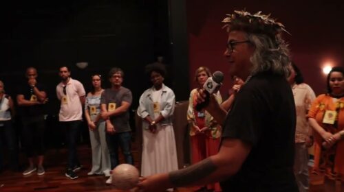 “Pessoa com cocar e microfone fala ao centro de uma roda de participantes em um auditório. O grupo está em pé, formando um círculo, enquanto a pessoa ao microfone segura um objeto arredondado nas mãos. Legenda em português aparece na parte inferior do vídeo.”