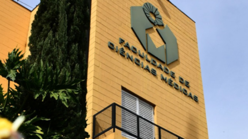 Prédio da Faculdade de Ciências Médicas da Unicamp, com fachada amarela, logotipo da instituição em destaque e jardim com flores em primeiro plano.