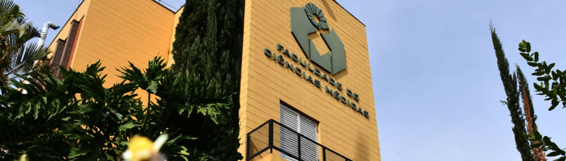 Prédio da Faculdade de Ciências Médicas da Unicamp, com fachada amarela, logotipo da instituição em destaque e jardim com flores em primeiro plano.