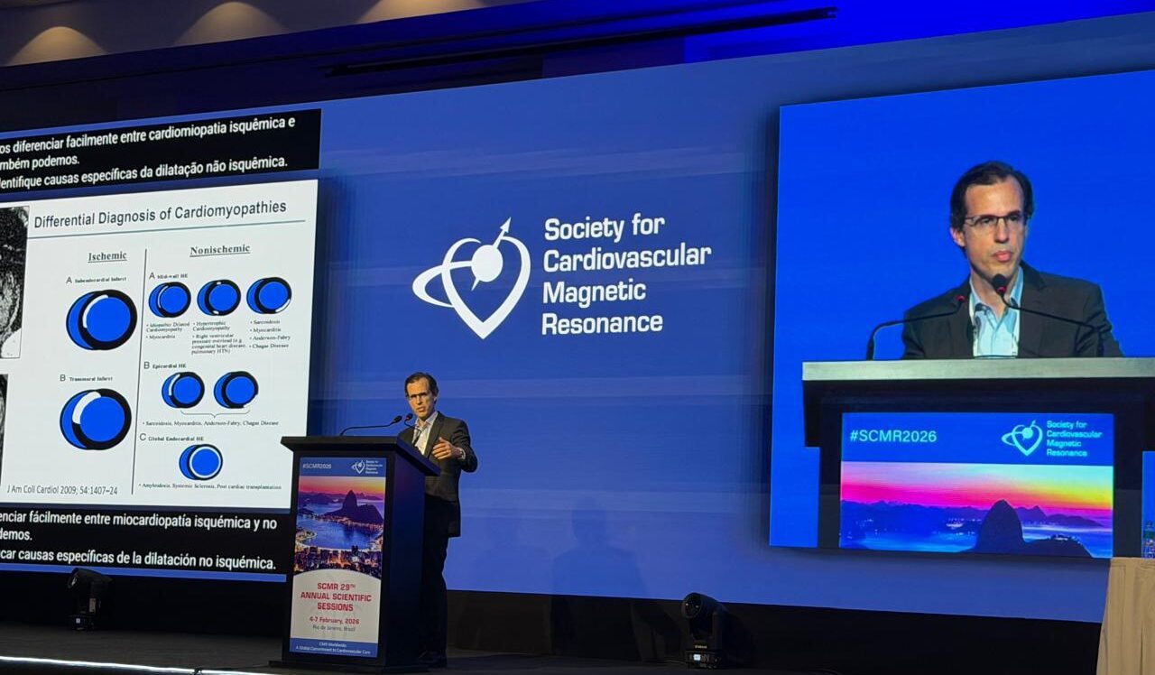 Auditório de congresso com iluminação azul. Um médico de terno escuro e óculos fala ao púlpito diante de uma grande tela com o logotipo da Society for Cardiovascular Magnetic Resonance. No telão lateral aparece um slide sobre diagnóstico diferencial de cardiomiopatias. No painel frontal do palco há a identificação “#SCMR2026” e uma imagem do Pão de Açúcar ao entardecer.
