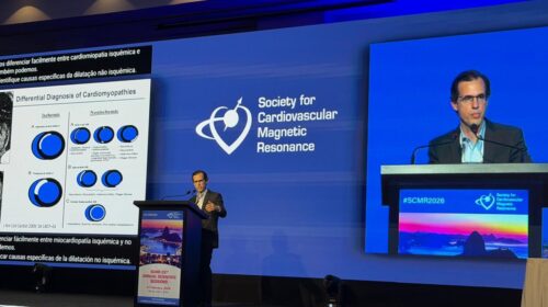 Auditório de congresso com iluminação azul. Um médico de terno escuro e óculos fala ao púlpito diante de uma grande tela com o logotipo da Society for Cardiovascular Magnetic Resonance. No telão lateral aparece um slide sobre diagnóstico diferencial de cardiomiopatias. No painel frontal do palco há a identificação “#SCMR2026” e uma imagem do Pão de Açúcar ao entardecer.