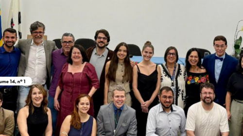 Foto de grupo em auditório institucional. Cerca de 25 a 30 pessoas posam em duas fileiras, algumas em pé e outras agachadas na frente, olhando para a câmera. Ao fundo há uma mesa de conferência, um telão branco e três bandeiras — do estado de São Paulo, do Brasil e da Unicamp. À direita da mesa há um vaso com orquídeas. O ambiente tem iluminação de teto e paredes em tons claros e verdes. Algumas pessoas seguram sacolas de papel, sugerindo participação em evento ou cerimônia.