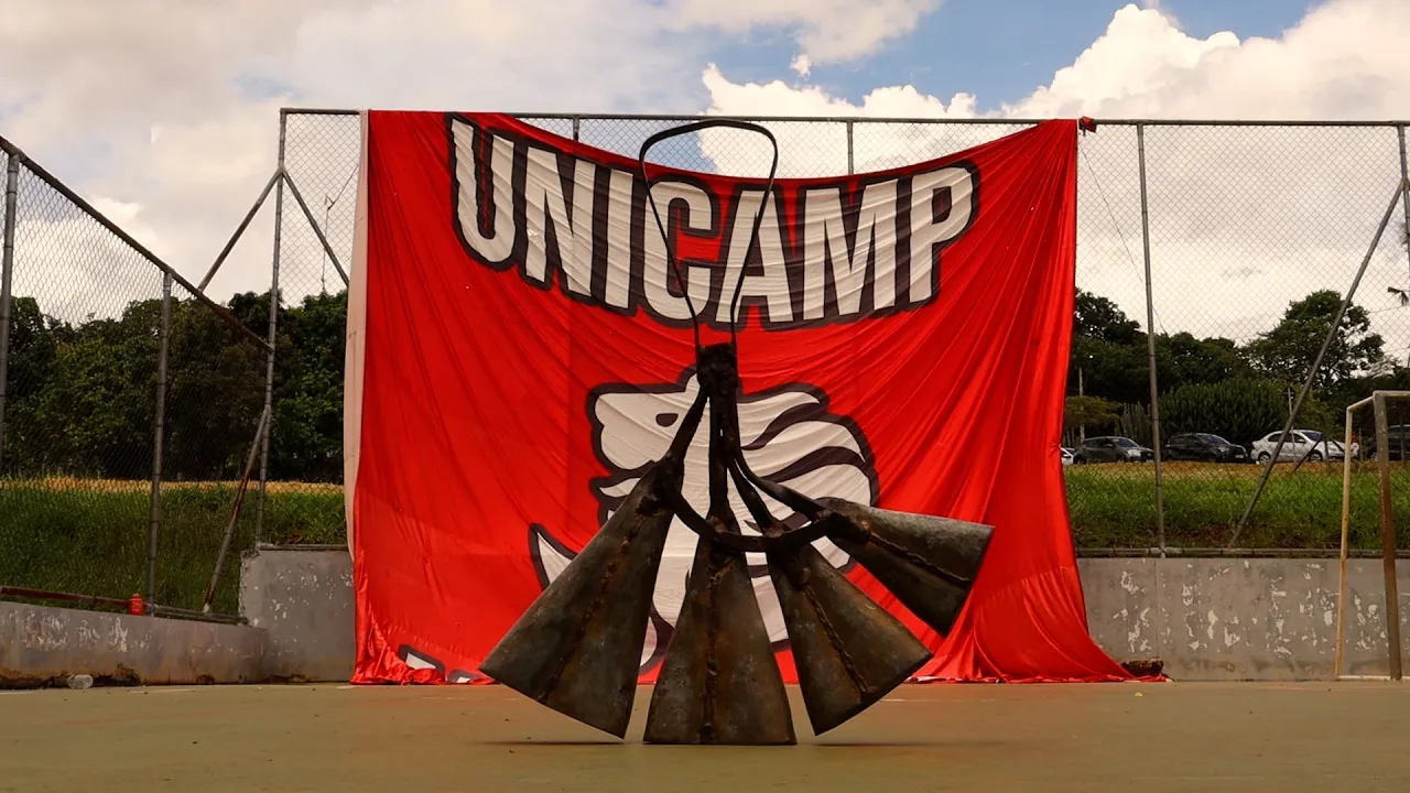 Escultura metálica com formato de pulmões em primeiro plano, instalada em uma quadra. Ao fundo, um grande bandeirão vermelho com a palavra “UNICAMP” preso a uma grade. O céu está parcialmente nublado e há vegetação e carros ao fundo.