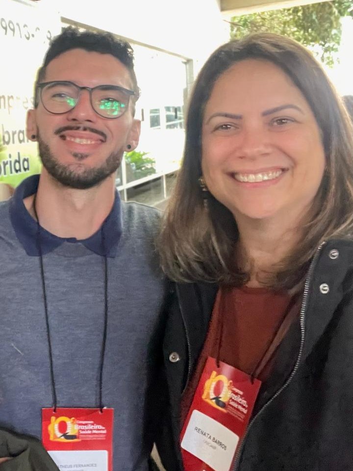 Selfie de plano medio de dos personas sonrientes. A la izquierda, un hombre con gafas, barba y un suéter gris con cuello azul. A la derecha, una mujer con cabello castaño y una chaqueta negra. Ambos llevan etiquetas rojas con sus nombres alrededor del cuello.